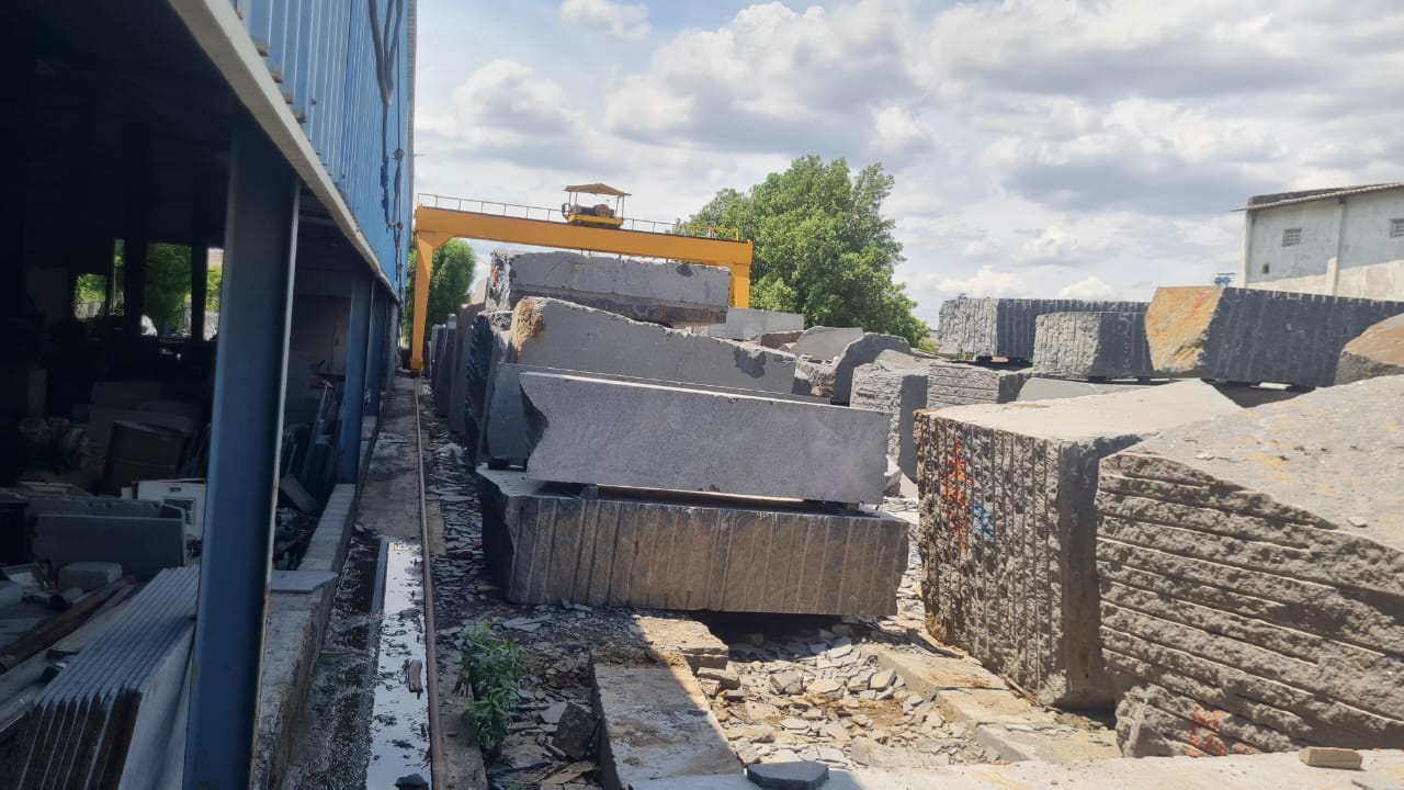 Rows of rough granite blocks in the yard beside the main shed, with a yellow gantry crane behind them